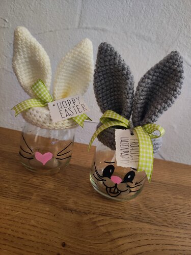 Zwei Gläser mit gehäkelten Hasenohren (weiß und grau), bemalten Gesichtern und grünen karierten Schleifen