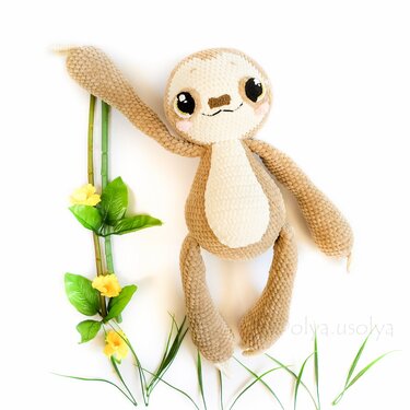 Beige crocheted amigurumi sloth toy with long arms lying next to green stems and yellow flowers