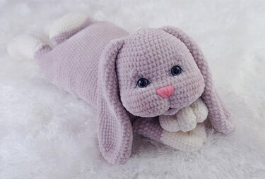 Lilac crocheted amigurumi rabbit lying on white fluffy fabric with long ears and pink nose