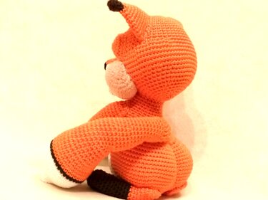 Side view of an orange crocheted amigurumi fox with white muzzle and black accents on a plain light background.