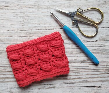 Coral-red bobble stitch crochet swatch beside a blue H (5.0 mm) crochet hook and ornate gold scissors on light wood.
