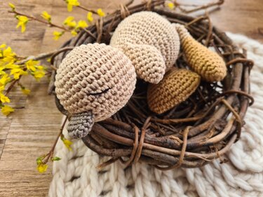 Beiges gehäkeltes Amigurumi-Küken mit geschlossenem Auge im Weidenkranznest neben gelben Forsythienzweigen