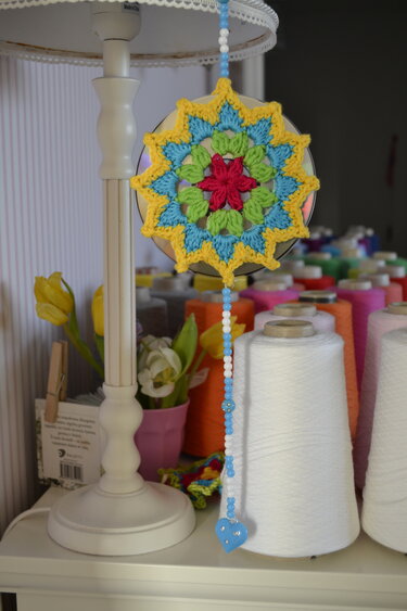 Gehäkeltes Mandala mit Perlenkette und blauem Herz vor Garnspulen