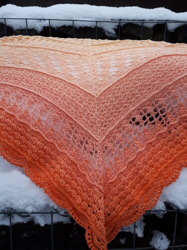 Gehäkeltes Dreieckstuch mit Orange‑Pfirsich-Farbverlauf und Spitzenmuster, auf einem schneebedeckten Geländer drapiert