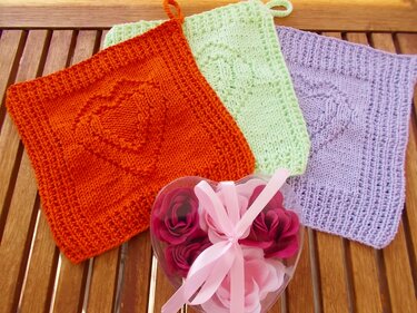 Drei gestrickte Topflappen (orange, hellgrün, flieder) mit Herzmotiv und Aufhängeschlaufen auf Holzuntergrund; herzförmige Geschenkbox mit Rosen daneben.