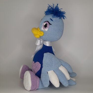 Gehäkeltes Amigurumi-Vögelchen in Hellblau und Dunkelblau mit gelbem Schnabel, großem Auge, weißer Schleife und lila Herz, sitzend vor hellem Hintergrund