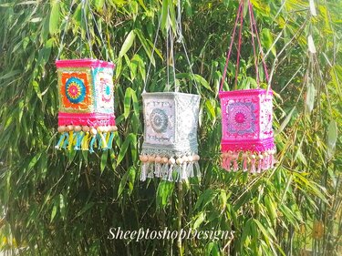 Three square crocheted lanterns in bright multicolor, pale green and pink, decorated with tassels and wooden beads, hanging against bamboo foliage.