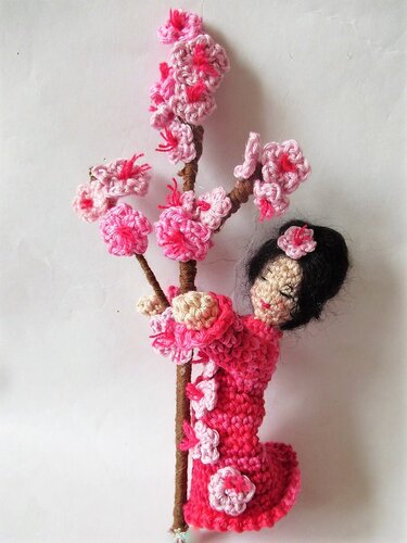 Gehäkelte Amigurumi-Puppe im rosa Kleid hält Zweig mit rosa Blüten vor weißem Hintergrund.