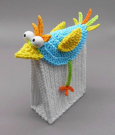 Colorful crocheted bird with oversized eyes and textured wings, mounted on a white textured crochet gift bag