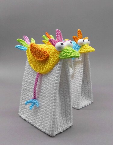 Two white crocheted rectangular gift bags with yellow bird-shaped crochet toppers, large white eyes and multicolored feather details.