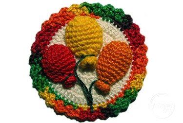 Runde gehäkelte Applikation mit drei plastischen Luftballons (Rot, Gelb, Orange) und mehrfarbigem Regenbogenrand auf weißem Hintergrund.