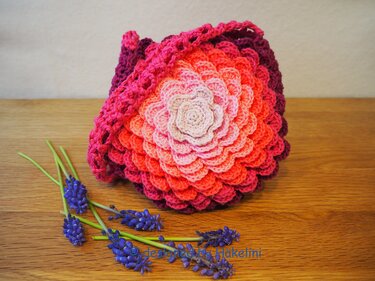 Handgehäkelte Tasche mit mehrschichtigem Blumenmotiv in Rosa- und Rottönen auf Holz, neben violetten Traubenhyazinthen.