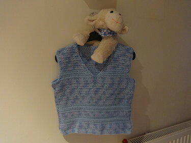 Hellblaue gehäkelte Kinderweste mit V-Ausschnitt an einem Kleiderbügel, darüber ein Stoffschaf