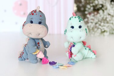 Two crocheted amigurumi dinosaurs, one blue and one mint, linked by a colorful crochet garland