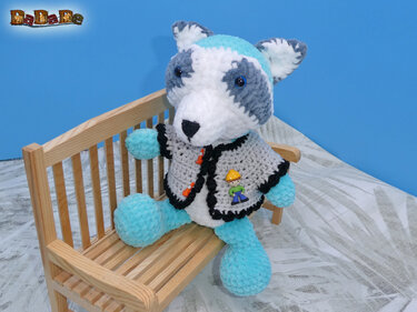 Gehäkelter Amigurumi-Teddy aus Plüschgarn in Blau und Grau mit Jäckchen auf Holzbank