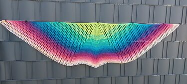 Halbrundes gehäkeltes Tuch mit buntem Farbverlauf an einem Zaun hängend