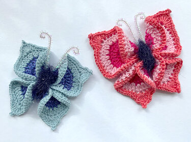 Zwei handgehäkelte Schmetterlinge in Rosa/Pink und Türkis/Blau mit flauschigem Körper und glänzenden Drahtfühlern auf hellem Hintergrund.