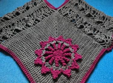 Grau gehäkelter Poncho mit pinker Blumenapplikation auf blauem Untergrund