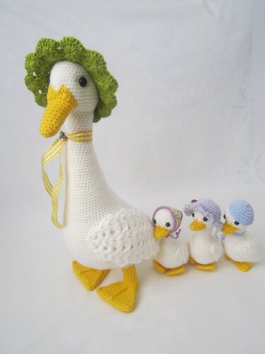 Weiße gehäkelte Gans mit grüner Rüschenmütze und drei kleinere Küken mit Pastellmützen auf weißem Hintergrund.