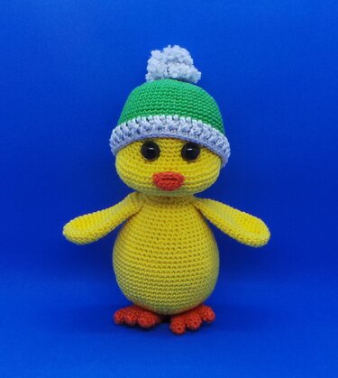 Gehäkeltes gelbes Küken mit grüner Bommelmütze auf blauem Hintergrund.