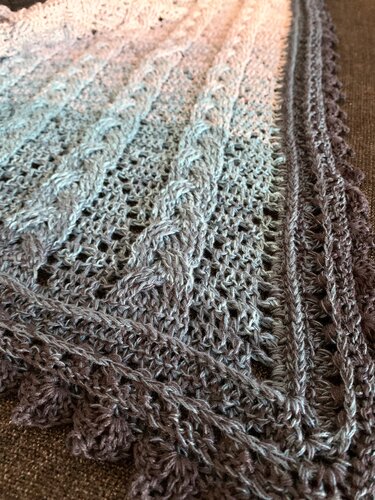 Close-up of a crocheted triangle shawl in gradient greys showing lace stitch patterns, cable panels and scalloped edging