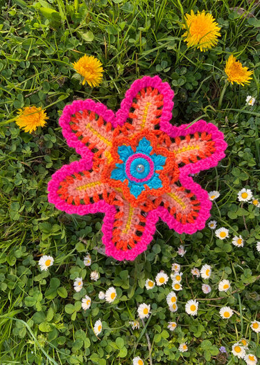 Bunte gehäkelte Blumenapplikation mit Zierknopf, liegend auf Gras mit Löwenzahn und Gänseblümchen.