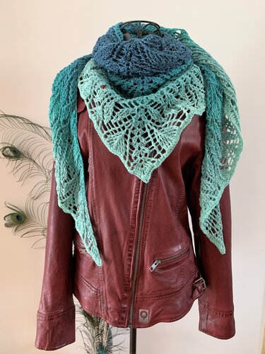 Ombre blue-to-green crocheted triangle shawl in an openwork lace pattern draped over a red leather jacket with peacock feathers nearby.
