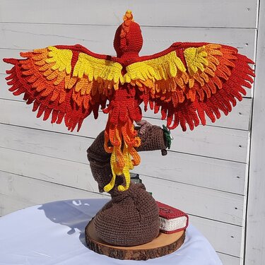Gehäkelter Phönix in Rot, Orange und Gelb mit ausgebreiteten Flügeln auf rundem Holzsockel