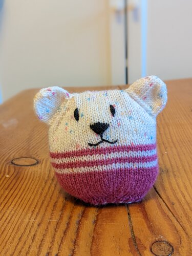 Gestricktes Amigurumi-Bärchen in Cremefarben und Magenta mit gesticktem Gesicht auf Holztisch