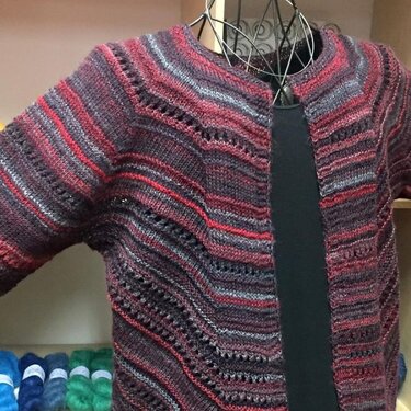 Striped knitted cardigan in red, purple, and gray with diagonal lace-stitch details on a mannequin.