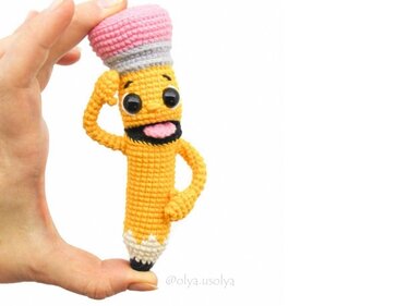 Hand holding a yellow crocheted pencil amigurumi with a smiling embroidered face and pink eraser on a white background.