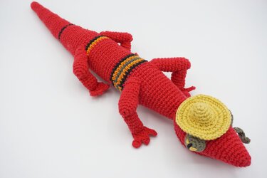 Red crocheted amigurumi lizard with multicolor stripes wearing a small yellow sombrero on a white background.