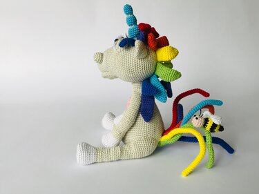 Seitliche Ansicht eines gehäkelten Amigurumi-Einhorns mit Regenbogenmähne und -schweif; am Schweif sitzt eine kleine gehäkelte Biene.