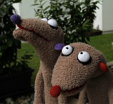 Zwei braune, flauschige Handpuppen mit großen Kulleraugen und farbigen Nasen vor grünem Hintergrund