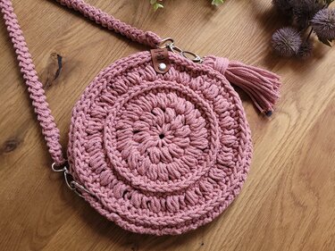 Soft pink round crochet bag with textured stitch motifs, braided strap and tassel laid on wood