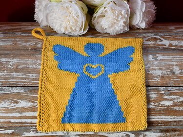 Quadratischer gelber Topflappen mit blauem Engelmotiv und Herz auf rustikalem Holztisch, oben wei Blumen.