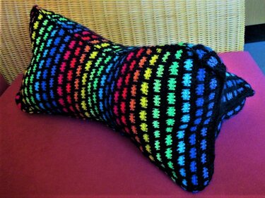 Längliches gehäkeltes Kissen mit buntem Regenbogen-Gittermuster auf schwarzem Grund, auf magentafarbenem Untergrund vor Korbgeflecht.