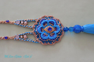 Nahaufnahme eines handgehäkelten Accessoires in Blau und Gold mit zentraler Blüte, blauem Schmuckstein, Perlen, Pailletten und einer blauen Quaste.