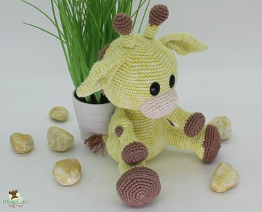 Hellgrüne gehäkelte Amigurumi-Giraffe mit braunen Akzenten, sitzend neben einer Topfpflanze und dekorativen Steinen.