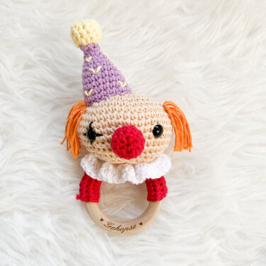 Gehäkelte Clown-Rassel mit lila Hütchen, orangefarbenen Zöpfen und roter Nase an einem Holzring mit Gravur 'Gehopse', auf weißem Kunstfell.