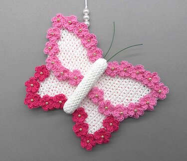 White crocheted butterfly with pink floral edging and gold-tone bead centers, wrapped white body and green wire antennae on a gray background.