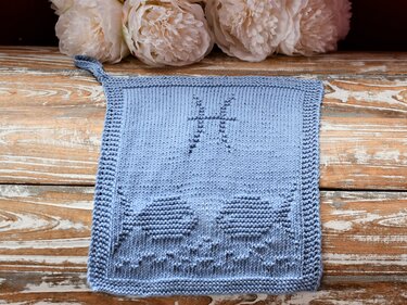Gestricktes hellblaues Quadrat mit eingearbeitetem Fische-Sternzeichen und zwei Fischformen auf rustikalem Holz