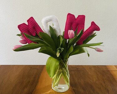 Gehäkelte MOM-Buchstaben in Pink und Weiß zwischen rosa Tulpen in einer Glasvase auf einem Holztisch