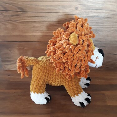 Gehäkelter Amigurumi-Löwe mit braun-orangefarbenem Körper, flauschiger orangefarbener Mähne und weißen Pfoten auf Holzuntergrund.