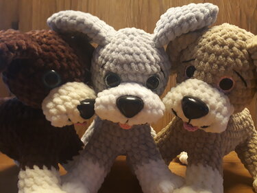 Drei gehäkelte Amigurumi-Hunde in braun, grau und beige mit weißen Schnauzen, runden Augen und schwarzen Nasen