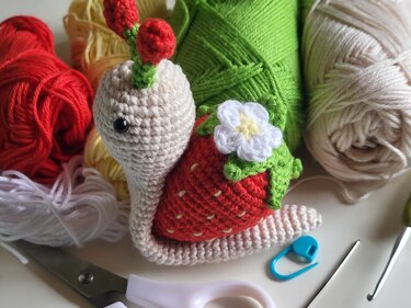 Amigurumi-Schnecke mit Erdbeerschale, umgeben von Garn, Häkelnadeln und Schere