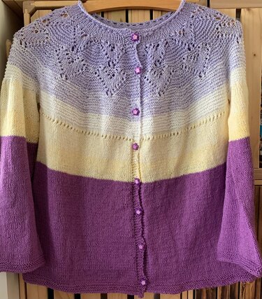 Gestrickte Kinderjacke mit Lochmuster am Rundkragen, breiten Streifen in Flieder, Hellgelb und Violett und lila Blütenknöpfen.