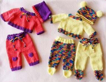 Flach ausgelegte, handgestrickte Puppenkleidung in Koralle, Gelb und bunten Mustern auf hellem Untergrund.