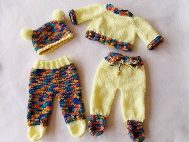 Vier gestrickte Teile für Puppen auf hellem Hintergrund: Mütze mit zwei Bommeln, Pullover sowie zwei Hosen mit farbigen Bündchen.