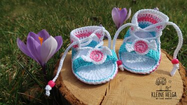 Gehäkelte Babysandalen in Weiß, Türkis und Rosa mit Blumenapplikation und Kordel auf Holzscheibe vor Gras und Krokussen